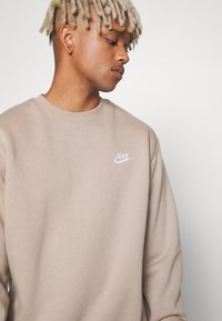 Bluza Nike w jasnym beżu, z klasycznym dekoltem, ozdobiona małym białym logo na piersi. Gładka tekstura, luźny krój.