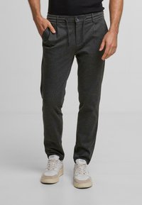 Pantalons de jogging gris avec chevilles resserrées, taille avec cordon de serrage et poches latérales, fabriqués en tissu doux ; associés à des baskets blanches et beiges.