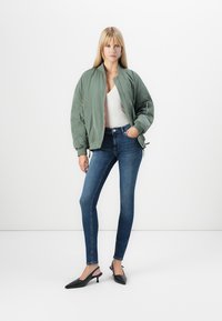 Olive green bomber jacket with ribbed collar, paired with a white top and dark blue skinny jeans. Black pointed-toe slingback heels.