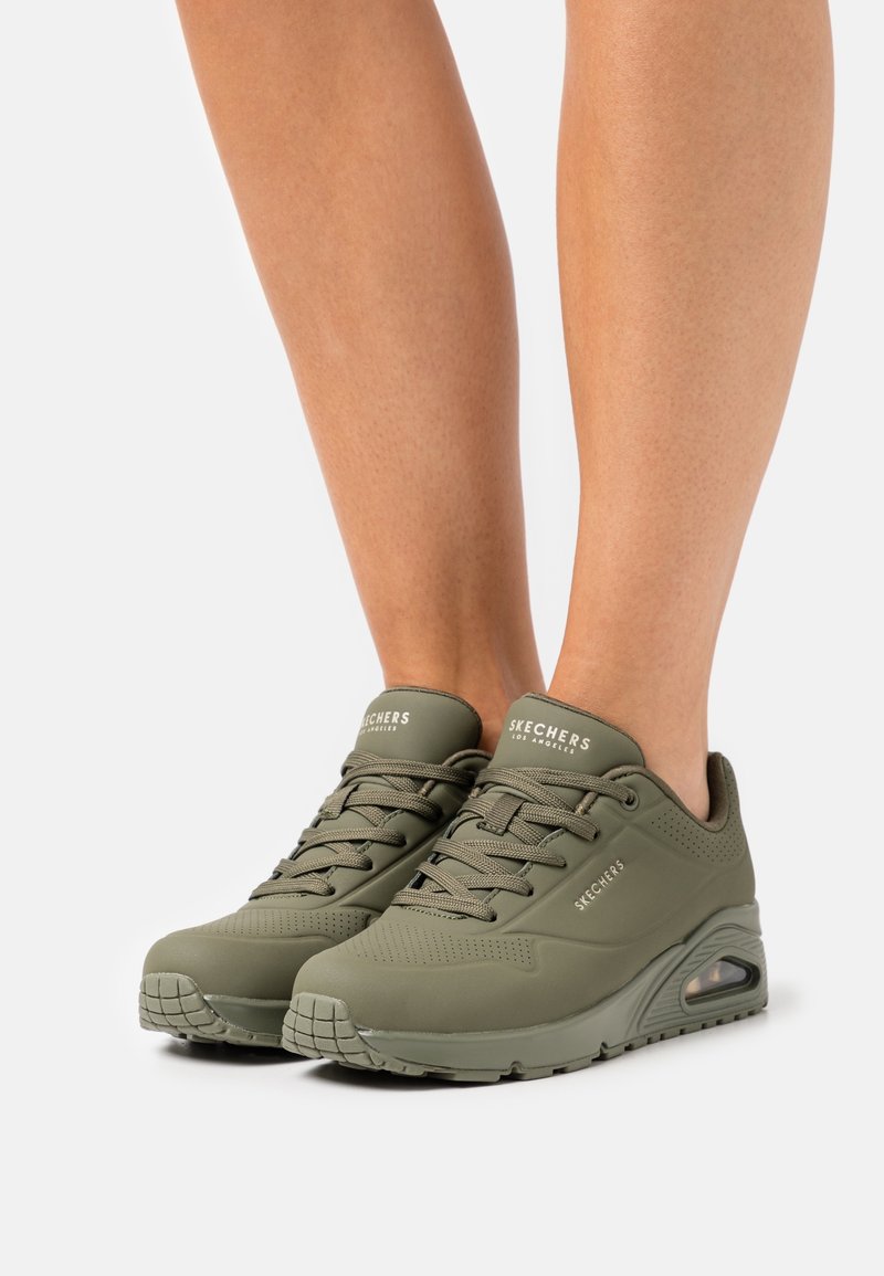 Personne portant des baskets Skechers vert olive avec un design à lacets et des semelles à coussin d'air visibles, debout sur un fond blanc.