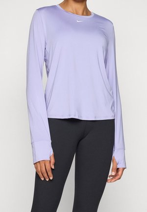 Frau trägt ein langärmliges Sportshirt in Lavendel mit Daumenlöchern und schwarze Leggings, steht vor einem schlichten Hintergrund.