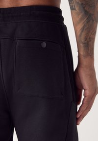 Zwarte sportbroek met een gestructureerde stof, voorzien van een achterzak die wordt afgesloten met een ronde knoop. Elastische tailleband voor comfort.