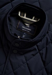 Granatowa pikowana kurtka z stojącym kołnierzem, dwoma czarnymi guzikami oraz metką z napisem "Matinique Heritage Apparel" wewnątrz.
