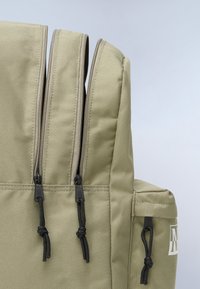 Zaino khaki con tre scomparti con cerniera, hardware nera e tessuto testurizzato. Anelli per accessori e logo sui lati.