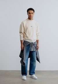 Jeune homme portant un pull crème, un jean bleu, une chemise à carreaux nouée autour de la taille et des baskets blanches, posé devant un fond uni.