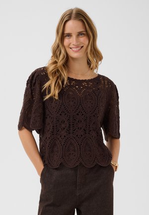 Femme souriante aux longs cheveux blonds portant un haut court à manches courtes en crochet marron foncé et un pantalon foncé, les mains dans les poches.