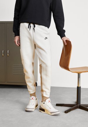 Persona con pantalones deportivos blancos Nike y zapatillas blancas Air Jordan junto a una silla de madera sobre un suelo gris.