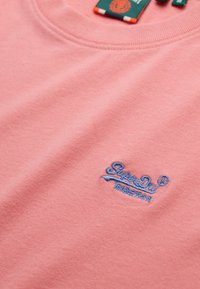 Roze crewneck sweatshirt met blauw geborduurd "SuperDry" logo en Japanse karakters op de borst, stoflabel zichtbaar bij de kraag.