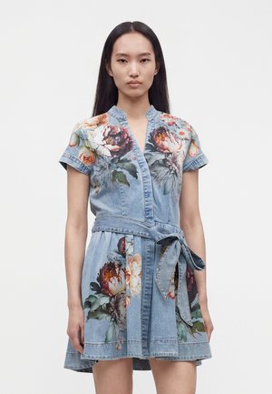 Femme portant une robe en denim bleu clair à manches courtes, imprimés floraux, ceinture nouée à la taille et col en V, debout devant un fond uni.