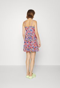 Vestido floral sem mangas com alças ajustáveis, padrão colorido de flores rosas, azuis e vermelhas, e saia rodada. Combinado com tamancos verdes.