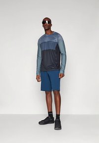 Αθλητικό μπλουζάκι με μακριά μανίκια σε γραδαρισμένα χρώματα ναυτικού μπλε και τυρκουάζ, συνδυασμένο με μπλε σορτς. Το ύφασμα φαίνεται λείο με εφαρμοστή σχεδίαση. Φοράει μαύρα αθλητικά παπούτσια.