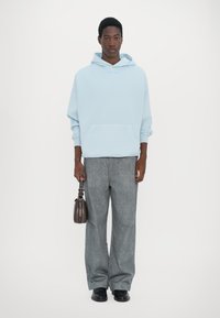 Felpa oversize azzurro chiaro con tasca frontale, pantaloni grigi a gamba larga e scarpe nere; tiene una piccola borsa marrone.