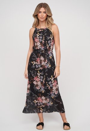 Giovane donna che indossa un vestito midi nero a fiori senza maniche e sandali neri slip-on, in piedi davanti a uno sfondo chiaro uniforme.