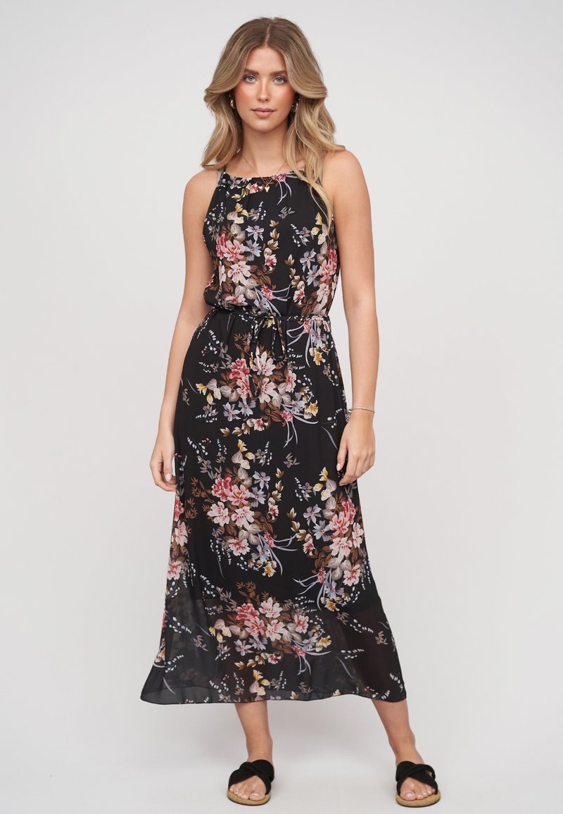Jeune femme portant une robe midi noire à fleurs sans manches et des sandales noires sans brides, debout devant un fond clair uni.