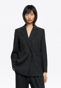 Blazer de cuadros oscuros con un solo botón, solapas con muescas, mangas anchas y bolsillos laterales. La tela texturizada añade profundidad al diseño.
