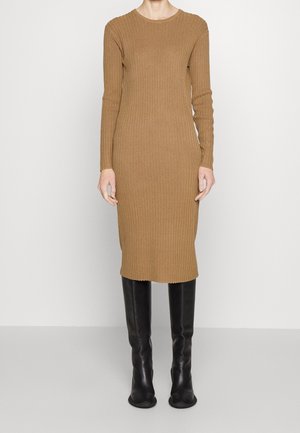 Kobieta w długiej, prążkowanej sukience w kolorze wielbłądzim, sięgającej do kolan, w połączeniu z czarnymi skórzanymi kozakami za kolano, stojąca na jednolitym tle.