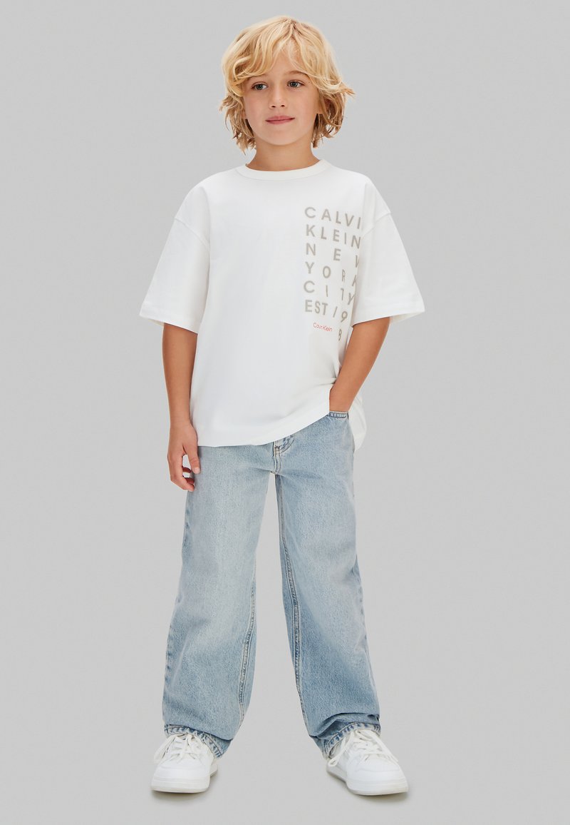 Blondes Kind steht mit einer Hand in der Tasche, trägt ein weißes Calvin Klein T-Shirt mit Grafik, hellblaue weite Jeans und weiße Turnschuhe.