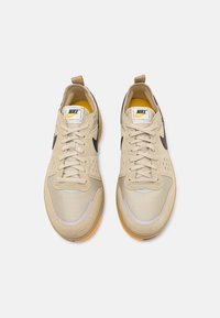 Ett par beige Nike-sneakers med svarta swoosh-loggor, mesh- och mockadetaljer, snören knutna, sedd uppifrån på en vit bakgrund.