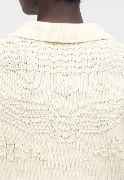 Πλάτη ατόμου που φοράει μπεζ μάλλινο πουλόβερ με ανάγλυφο έμβλημα φτερού και γεωμετρικά σχέδια στο πάνω μέρος της πλάτης.