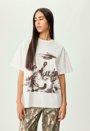 T-shirt en coton blanc avec un graphique marron d'un chat et d'un OVNI dans un paysage désertique. Manches courtes, coupe décontractée et encolure ras du cou.
