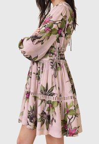 Femme portant une robe fleurie rose clair avec de longues manches bouffantes et une bordure en dentelle, nouée à la taille avec un nœud coulissant.