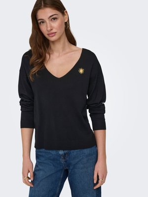 Femme aux longs cheveux bruns portant des boucles d'oreilles créoles en or, un pull noir à col en V avec un petit emblème doré en forme de soleil, et un jean bleu.