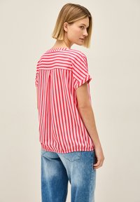 Rød og hvid stribet kortærmet bluse med en samlet kant og rygdetalje, lavet af letvægtsstof, parret med blå jeans.