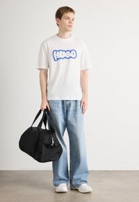 Hvid T-shirt med blå "HUGO" tekst, løse lyseblå jeans og sort sportstaske, parret med hvide sneakers på en grå baggrund.