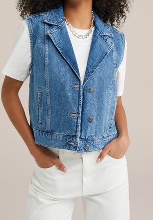 Persona que lleva un chaleco sin mangas azul de mezclilla sobre una camisa blanca de manga corta, pantalones blancos y un collar de cadena plateada.