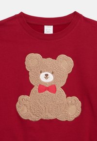 Red children's shirt with a textured teddy bear patch wearing a red bow tie centered on the front.