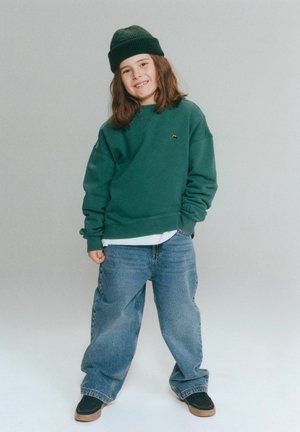 Uśmiechnięte dziecko w zielonej czapce i bluzie, szerokich dżinsach oraz czarnych butach stoi na jednolitym jasnoszarym tle.