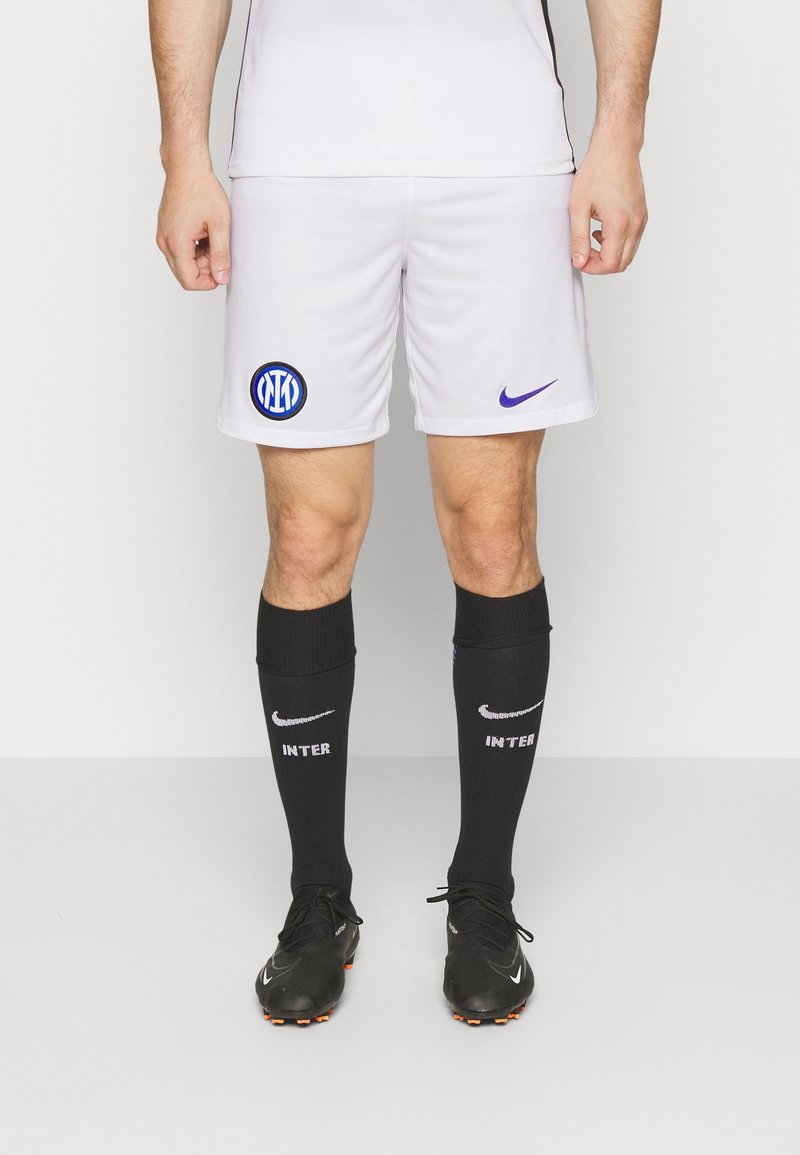 Athlète masculin portant un short de football blanc de l'Inter Milan, des chaussettes noires montant jusqu'aux genoux avec des logos Nike, et des crampons noirs, debout sur un fond blanc.