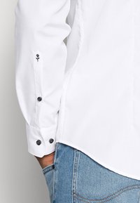 Valkoinen pitkähihaisessa paidassa, jonka hihansuut ovat mustat napit ja pienet mustat kukka-embroideringit, yhdistetty sinisiin farkkuihin, käsi taskussa.