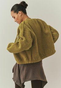 Ženska s temnimi lasmi v figi, oblečena v prevelik teksturiran olivno zelen jopič in rjavo asimetrično krilo na enobarvnem ozadju.