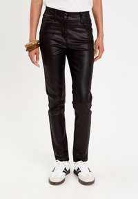 Pantalones de imitación de cuero negros con un ajuste slim, cintura alta, cierre de dos botones, bolsillos frontales y una textura suave y brillante. Usados con zapatillas blancas.