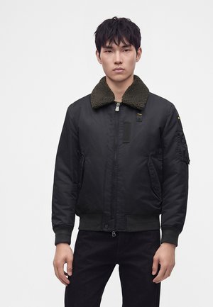 Jeune homme portant une veste bomber noire avec un col sherpa vert foncé, debout devant un fond blanc uni avec une expression neutre.