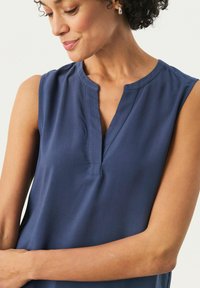 Marineblaue ärmellose Bluse mit einem Reverskragen, subtiler sheen und glattem Stoff. Sie hat eine lockere Passform und kleine Nahtdetails am Kragen.