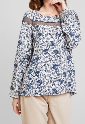 Woman wearing a long-sleeve white blouse with blue floral print and sheer yoke detail, paired with light beige pants.