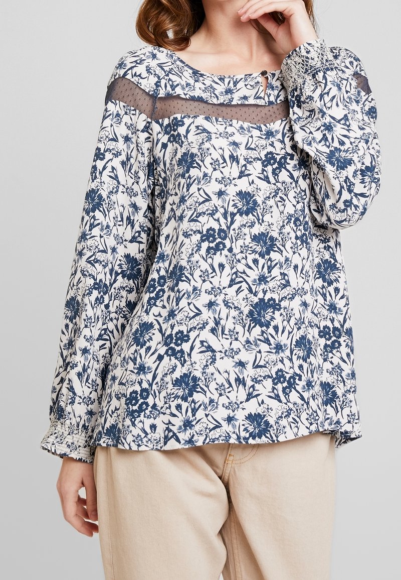 Femme portant un chemisier blanc à manches longues avec un imprimé floral bleu et un détail en tulle transparent sur le haut, associé à un pantalon beige clair.
