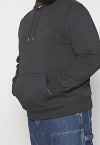 Sweat à capuche gris foncé en mélange de coton, avec une poche avant, des poignets côtelés et un petit emblème carré sur la poitrine.