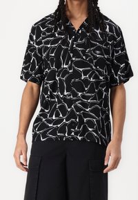 Černé tričko s krátkým rukávem s bílým abstraktním vzorem, vyrobené z měkké látky, s designem na zapínání na knoflíky a jemným detailem loga.