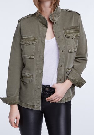 Persoon die een olijfgroen jasje met knopen draagt over een wit overhemd en zwarte leren broek, met één hand bij de taille.