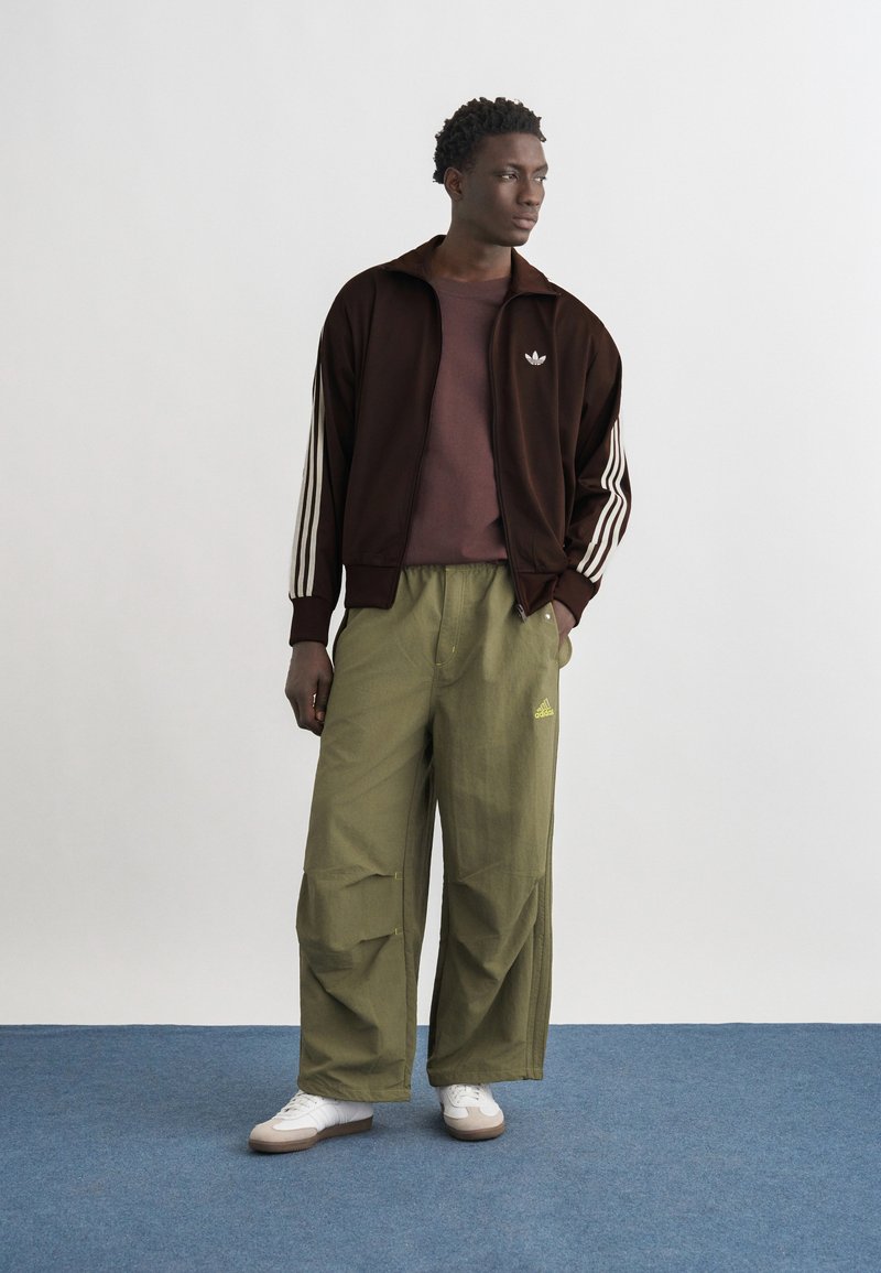 Uomo in piedi su un tappeto blu che indossa una giacca sportiva Adidas marrone, una maglietta color prugna, pantaloni larghi verde oliva con logo, e sneaker bianche.