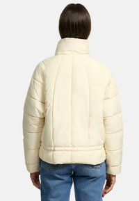 Cream puffed jacket with a high collar, ribbed texture, and visible stitching. Worn over blue jeans, featuring a cropped design and subtle branding.
