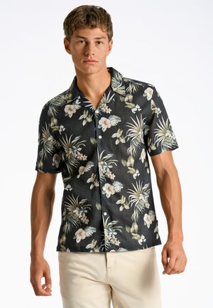 Camisa de manga corta, de fondo oscuro con flores en negro, con hojas verdes y flores blancas, con cuello y botones en la parte delantera. Tejido ligero.