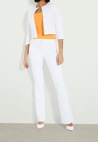 Veste en denim blanc à manches trois-quarts avec des boutons argentés, associée à un débardeur orange ajusté et un jean évasé blanc avec une finition texturée.