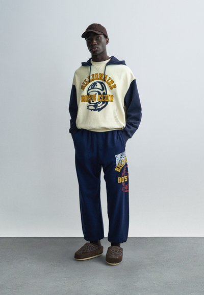 Φούτερ με κουκούλα σε εκρού και ναυτικό μπλε με γραφικά "Billionaire Boys Club", συνδυασμένο με ναυτικό μπλε φόρμα με τυπωμένα σχέδια και καφέ παντόφλες.