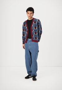Cardigan cu un model albastru și maroon, cu forme geometrice, cu închidere cu nasturi și manșete ribbate. Asortat cu blugi casual de un albastru deschis.
