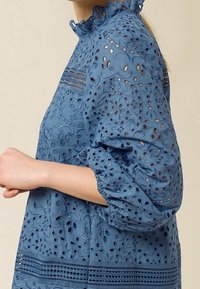 Blusa azul bordada con un escote alto y con volantes, mangas abullonadas, intrincados patrones florales y acentos de rayas horizontales.