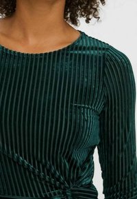 Top de terciopelo verde oscuro, con rayas verticales, mangas largas y un detalle anudado en la cintura. La textura suave realza el diseño.